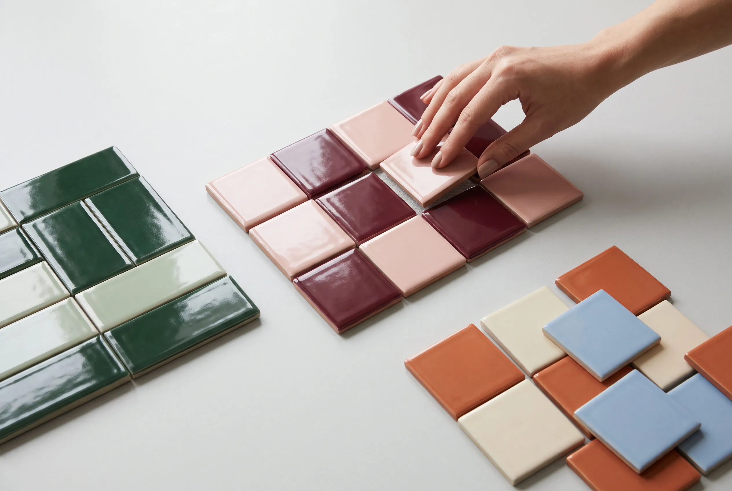 Glossy ceramic tiles in forest green, sage, blush pink, burgundy and terracotta arranged in geometric patterns on a grey surface, with a hand placing one tile